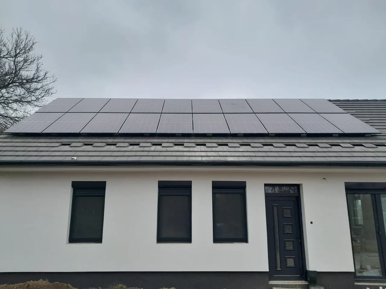 A képen egy modern, fehér falú ház látható, amelynek tetején napelemek sorakoznak. A napelemek fényes felülete jól látható a szürke égbolt alatt. A ház bejárati ajtajának kerete sötét színű, és a nyílászárók is modern stílust képviselnek. A napelem telepítés Dócon, az Ares Solar Construction Kft. szakértelmével készült, hangsúlyozva a fenntartható energiahasználat fontosságát.