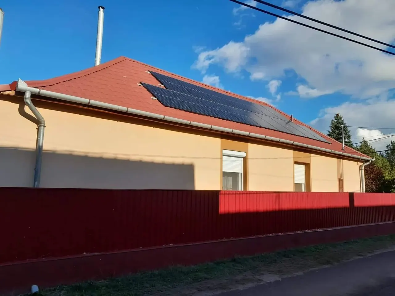 A képen a Pécs városában található Ares Solar Construction Kft. napelem telepítése látható. A tetőn több napelem sorakozik, melyek fényes felületei jól tükrözik a napfényt. A napelemek egy piros cseréptetőre vannak felszerelve, és a háttérben láthatóak a környező épületek, zöld fák és egyéb tetők, amelyek hozzájárulnak a városi környezethez.