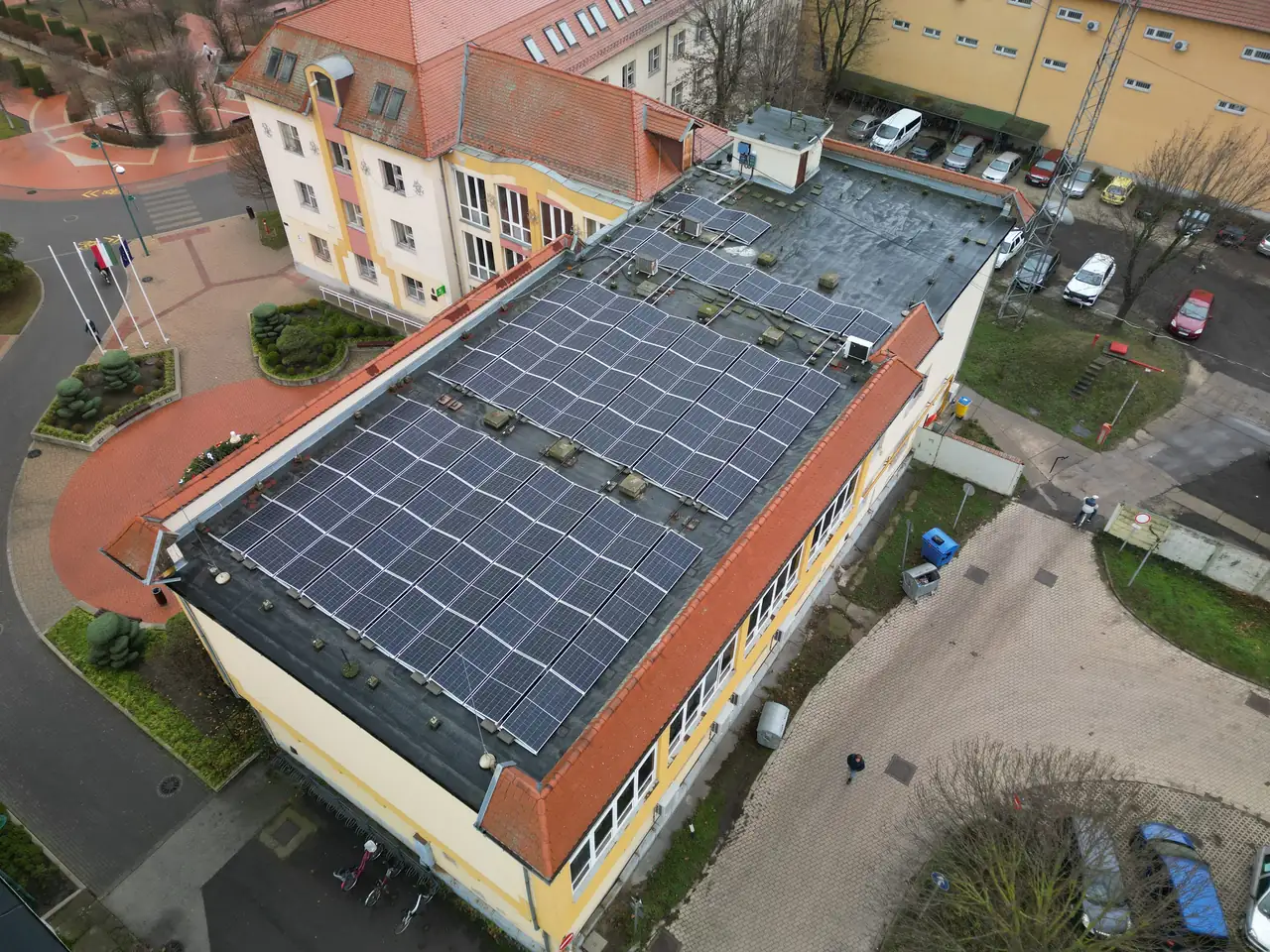 Dóc városában található Ares Solar Construction Kft. napelem telepítési projektje, amely egy épület tetején elhelyezett napelemeket mutat be. A képen jól láthatóak a fekete napelem panelek, amelyek a tetőn helyezkednek el, valamint az épület körüli terület, ahol parkolók és zöld területek találhatóak. Az épület modern megjelenése a napelemek által fenntarthatóbb energiatermelést képvisel.