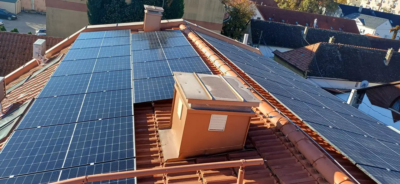 A képen egy tető látható, amelyen napelemek sorakoznak, napfényben csillognak. A napelemek a Tatabányai Ares Solar Construction Kft. napelem telepítési munkálatainak részei. A háttérben városi épületek és zöld fák láthatók, amelyek a város élénk hangulatát tükrözik. Az épület tetején egy szellőző is megfigyelhető, ami a tetőteret árnyékolja.