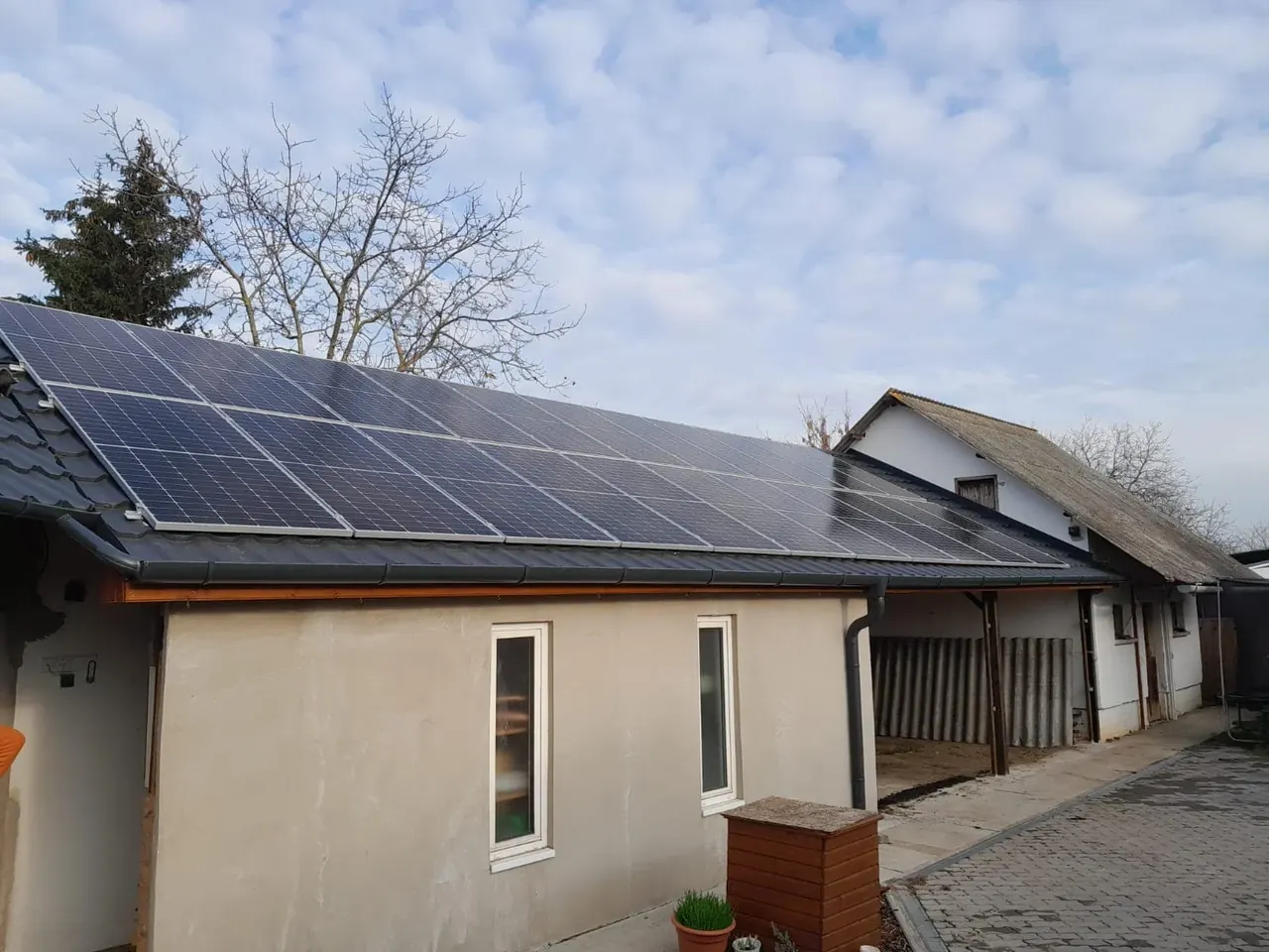 Dócon, az Ares Solar Construction Kft. napelem telepítési projektje látható egy ház tetején. A képen a modern napelemek feltűnően elhelyezkednek a szürke falú épület tetején, a háttérben fák és egy másik épület látható. A világos égbolt és a felhők növelik a napenergia hasznosításának érzését, tükrözve a cég elkötelezettségét a fenntartható energia iránt.