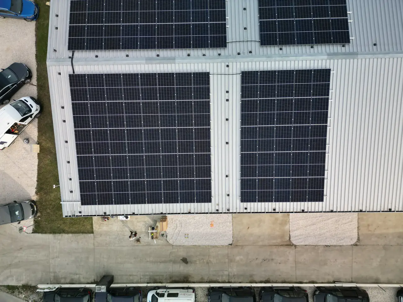 A képen a Salgótarjánban található Ares Solar Construction Kft. napelem telepítési projektje látható. A tetőn fekvő napelemek sorai szépen elhelyezkednek, míg a földszinten parkoló autók és a telepítési munkálatok során használt eszközök is láthatóak. A kép magasságából jól látszik a modern napelem rendszer, amely a fenntartható energia elősegítését szolgálja.