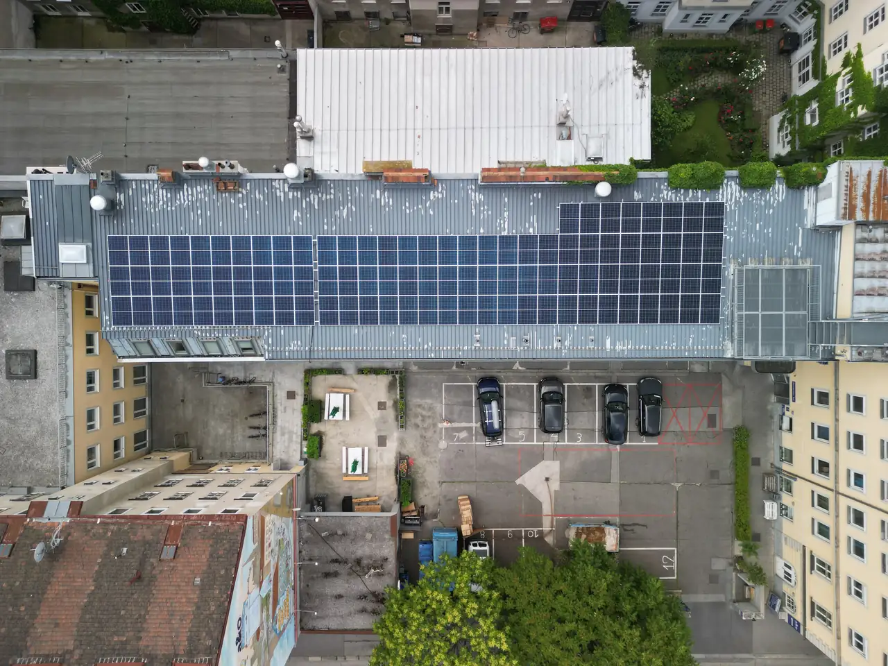 Dóc városában található Ares Solar Construction Kft. napelem telepítési projektjének légifelvétele, amelyen egy ház tetején elhelyezett napelemes rendszer látható. A napelemek szépen sorakoznak a fém tetőn, míg alatta parkoló autók és zöld területek találhatók. A felvételen jól látható a napelem telepítés hatékonysága és a városi környezet.