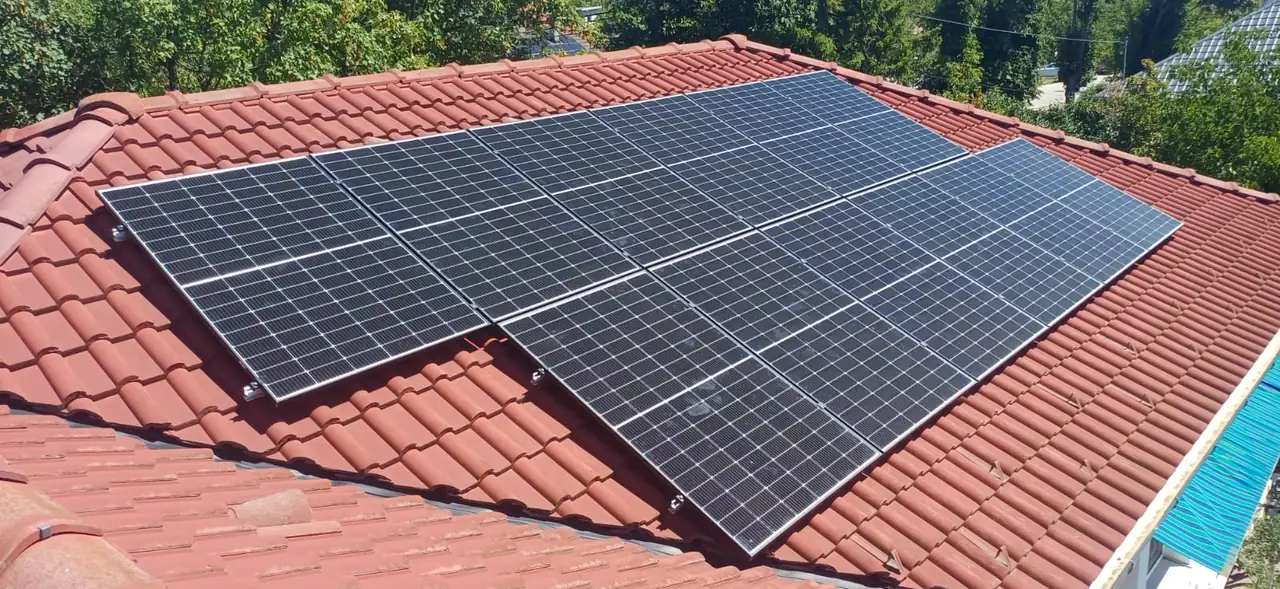 A képen egy tetőn elhelyezett napelem-rendszer látható, amelyet az Ares Solar Construction Kft. telepített Pécs városában. A napelemek sötét színűek, és jól láthatóan a cseréptetőre vannak rögzítve, maximálisan kihasználva a napfényt. A zöld környezet és a fák a háttérben jelzik a fenntartható energiaforrások iránti elkötelezettséget, amely a Napelem Telepítés területén kiemelkedő jelentőséggel bír.