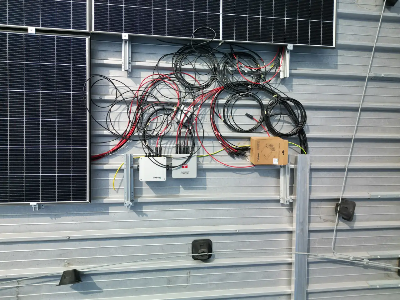Napelemek és elektromos vezetékek összekötött rendszere látható a képen, amely az Ares Solar Construction Kft. Szekszárdi telephelyén készült. A napelem telepítés során a különböző kábelek és inverterek precíz elhelyezése kulcsfontosságú a hatékony energiahasználat érdekében. A kép jól szemlélteti a szakmai munka részleteit és a napelem rendszer kiépítésének fontosságát.