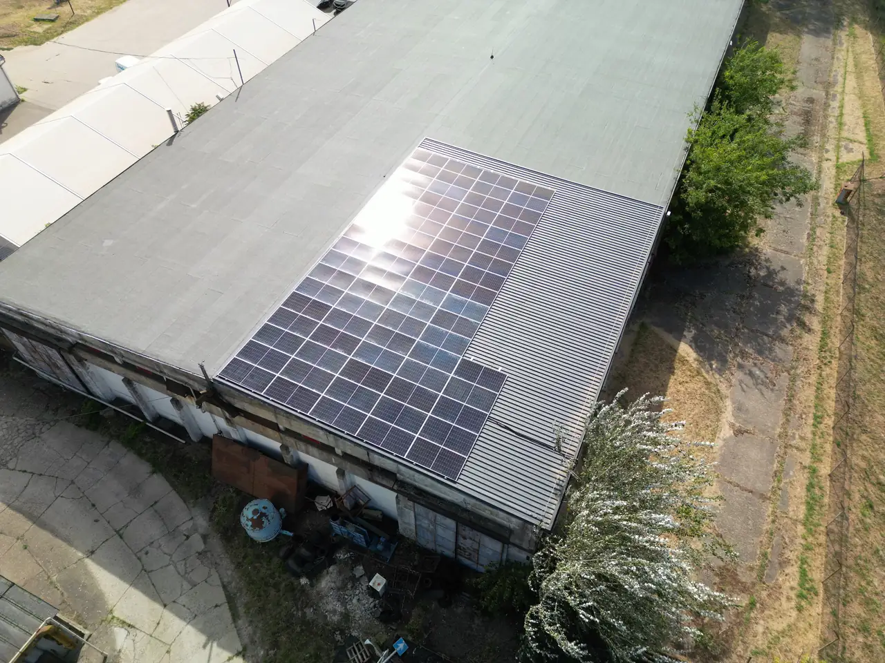 A képen egy Dócon található ipari épület tetőjén elhelyezett napelem rendszer látható, amelyet az Ares Solar Construction Kft. telepített. A napelemek szakszerűen vannak felszerelve, és tükrözik a napfényt, ami a környezet zöld energiával való ellátásának fontosságát hangsúlyozza. A képen a tető szerkezete és a környező terület is jól látható, ami a napelem telepítés helyét mutatja.