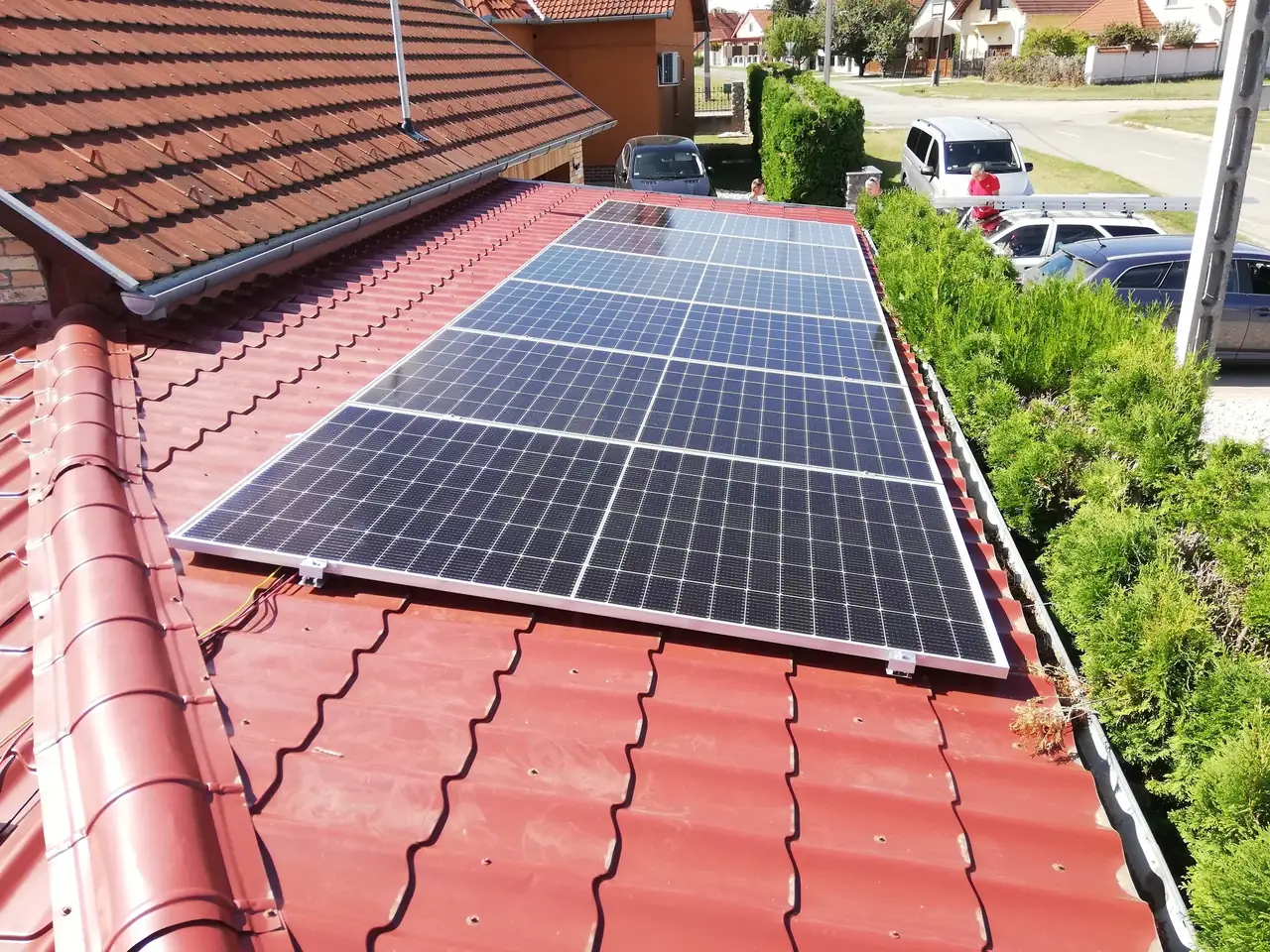 Napelemek a Dócon található ház tetőjén, a Ares Solar Construction Kft. által telepítve. A képen jól látható a piros cserép tetőszerkezet, amelyre a napenergia hasznosítására szolgáló napelemeket helyeztek el. A zöld növényzet és a környező épületek is láthatók, amelyek a modern napelem telepítés előnyeit hangsúlyozzák.