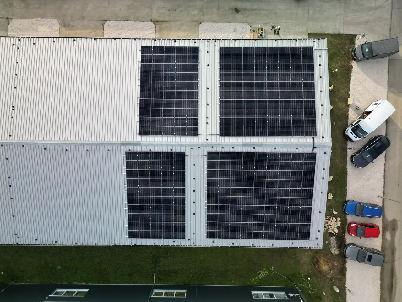 A képen a Dóc városában található Ares Solar Construction Kft. napelem telepítési projektje látható. A felvétel egy ipari létesítmény tetejéről készült, ahol négy napelem panel sorakozik. Az épület körül parkoló autók és zöld területek láthatók, amelyek hozzájárulnak a modern környezetbarát energiahasználathoz. A napelemek fényes felülete a napfényben ragyog, hangsúlyozva a fenntartható energia iránti elkötelezettséget.