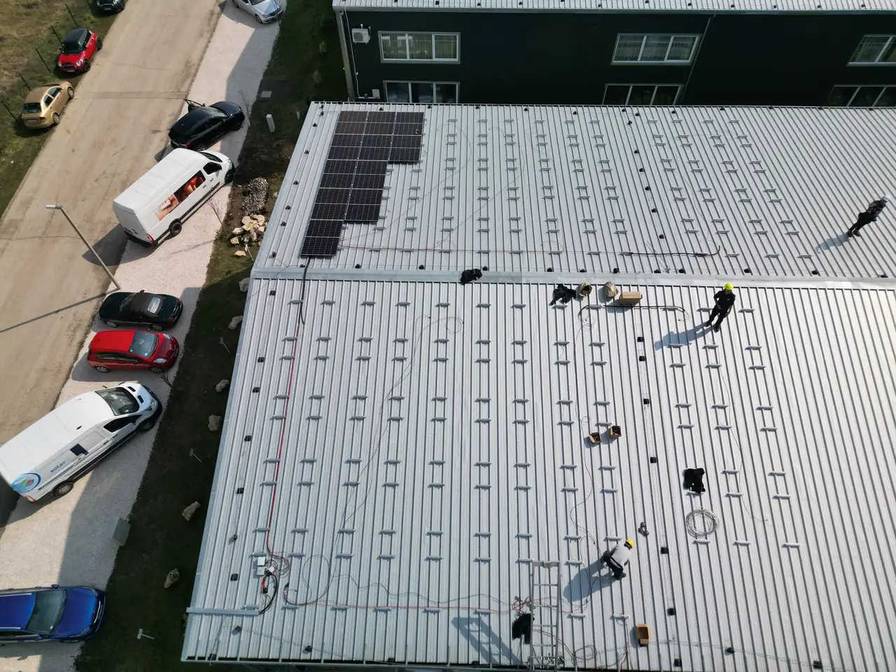 Dóc városában az Ares Solar Construction Kft. napelem telepítési projektje látható, ahol a szakemberek aktívan dolgoznak egy nagyobb épület tetőjén. A képen napelemek sorakoznak a fényes, fémes felületen, miközben a munkások különböző pozíciókban készítik elő a telepítést. Az autók a környező területen parkolnak, ami a munka intenzitását tükrözi.