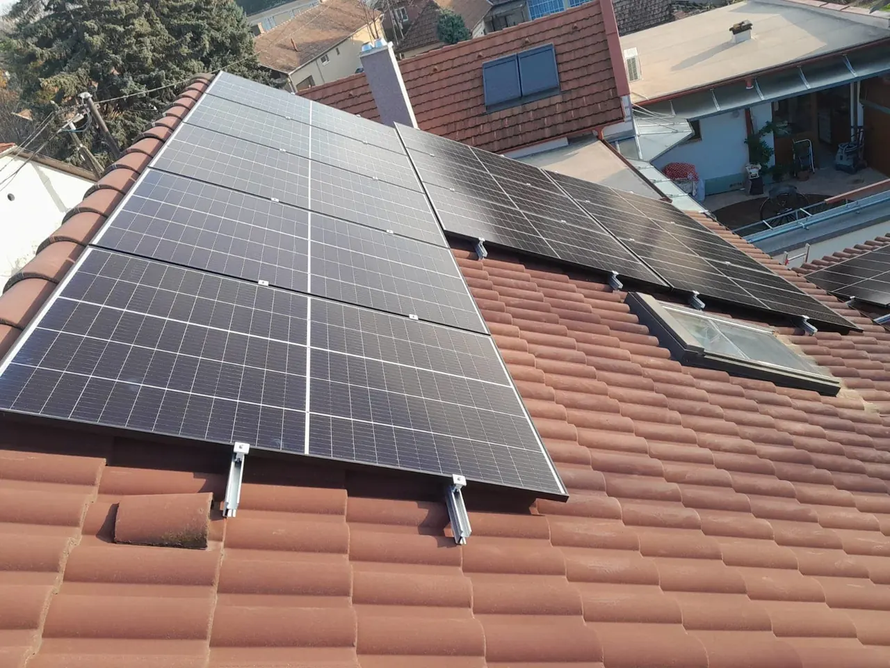A képen egy ház tetőjén elhelyezett napelemes panelek láthatók, amelyek a Dóc településen található Ares Solar Construction Kft. Napelem Telepítés szolgáltatásának részeként lettek telepítve. A paneleket jól láthatóan rögzítették a cseréptetőre, ami napos időben kiváló energiafelhasználást ígér. A háttérben további házak és a város nyugodt környezete is megfigyelhető.