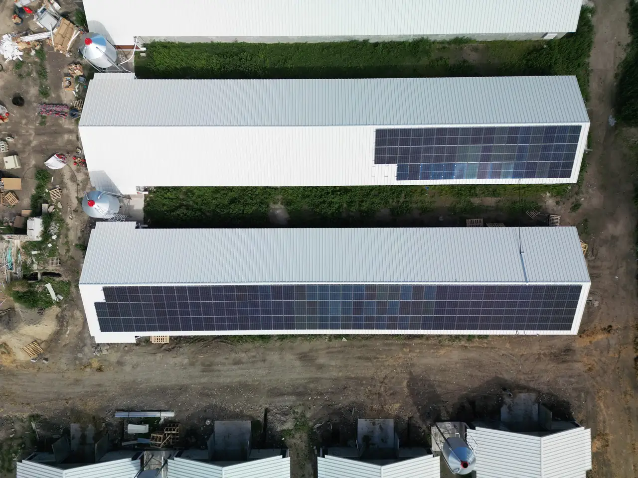A képen Dóc városában található napelem telepítés látható, ahol az Ares Solar Construction Kft. munkatársai dolgoznak. A felvételen három hosszú épület tetején elhelyezett napelemek sorakoznak, amelyek hatékonyan hasznosítják a napenergia erejét. A telepítés környezetét zöld növényzet és építőipari gépek övezik, ami a napelemek telepítésének aktív fázisát jelzi.