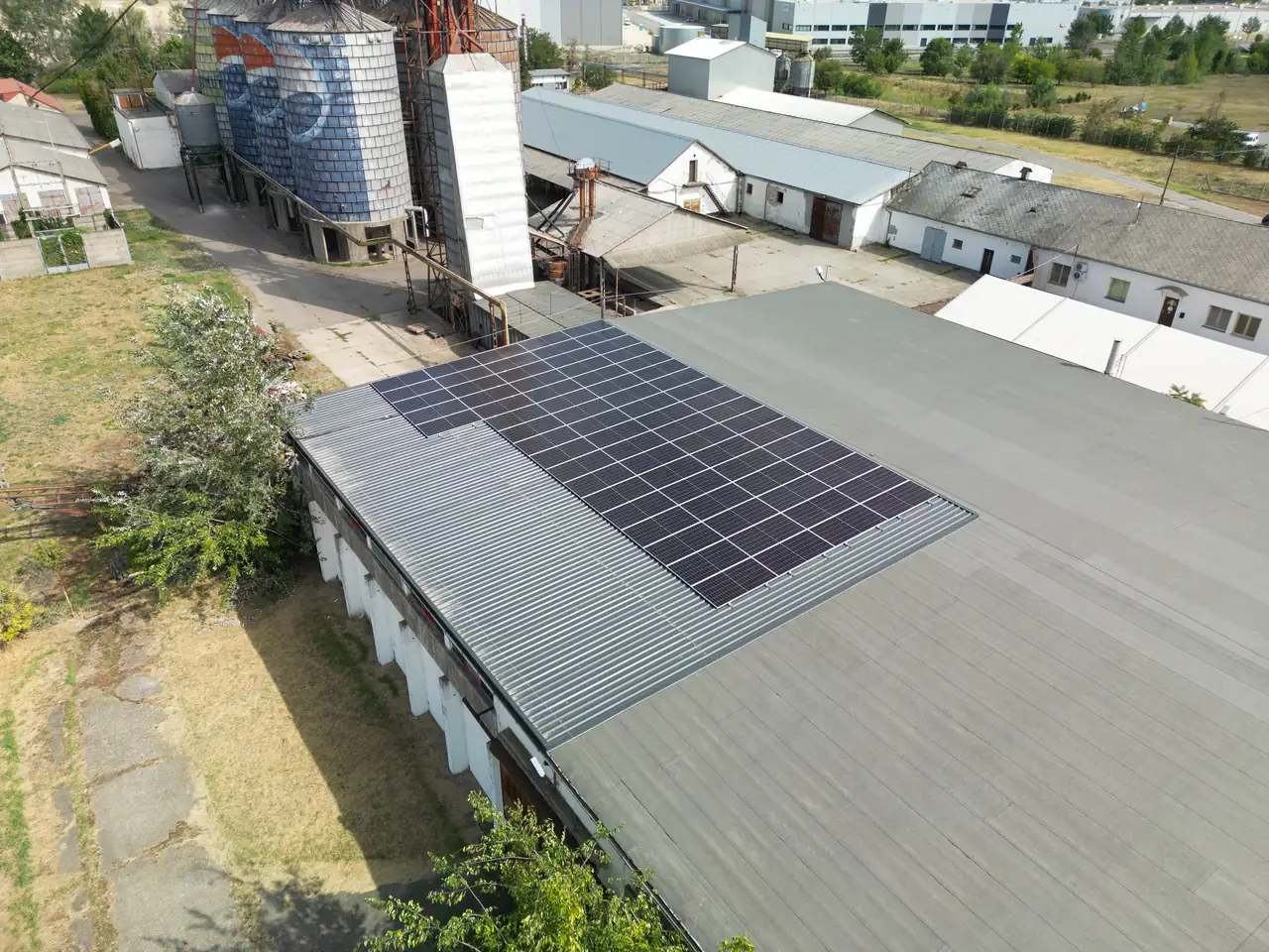 A képen egy Dócon található ipari épület tetején elhelyezett napelem rendszer látható, amelyet az Ares Solar Construction Kft. telepített. A napelemek jól láthatóan szögletes formájúak, és a tető szürke borításán helyezkednek el. A háttérben több épület és ipari berendezés is megfigyelhető, ami a napelem telepítés környezetét mutatja be.