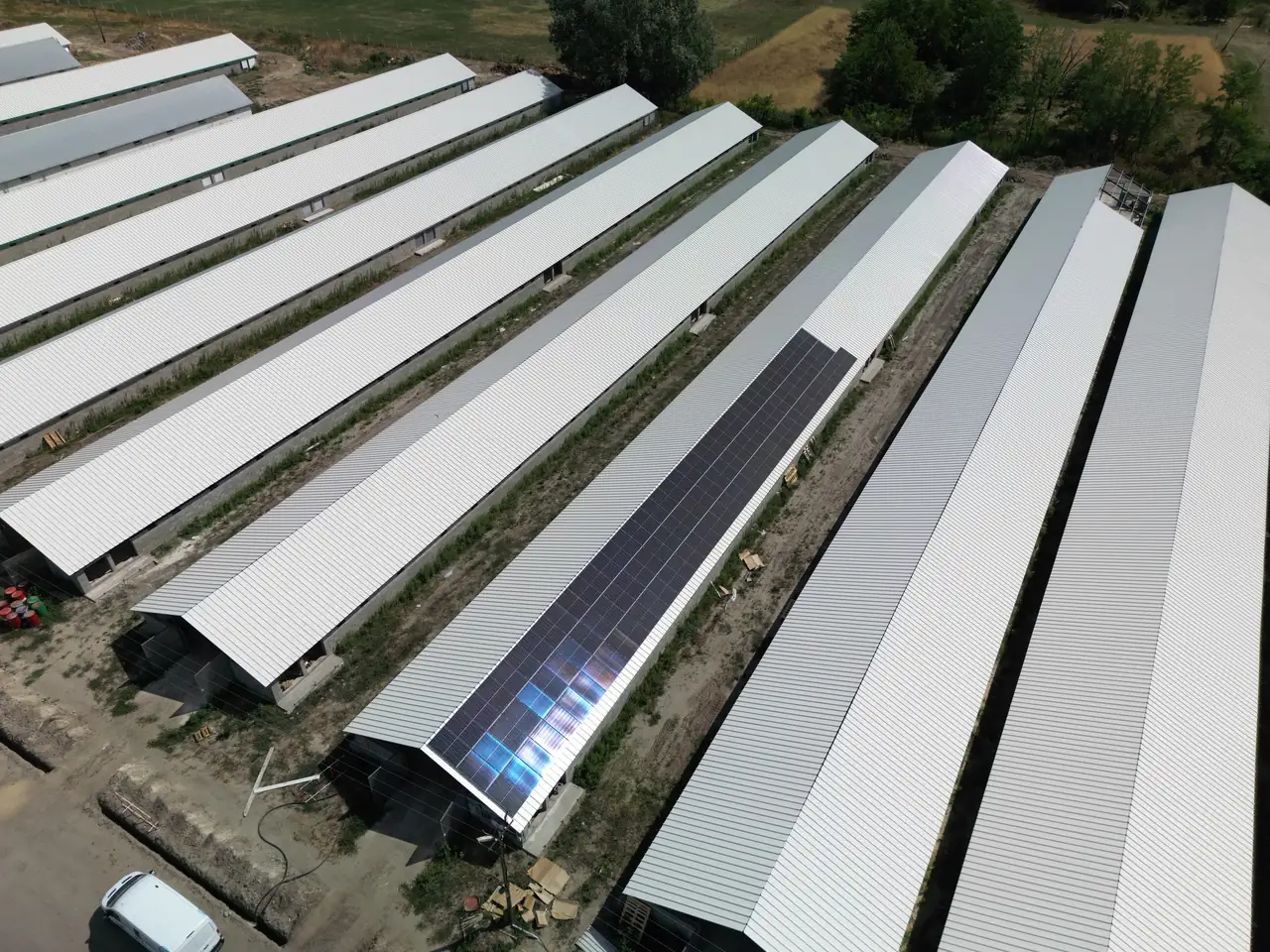 A képen Dóc városában található napelem telepítés látható, ahol a Ares Solar Construction Kft. munkatársai dolgoznak. Az épületek tetején sorakozó napelemek fényesen csillognak a napfényben, míg a háttérben zöldellő fák és mezők láthatók. A telepítés hatékonyan kihasználja a tetők felületét, biztosítva a megújuló energiaforrások maximális kihasználását.
