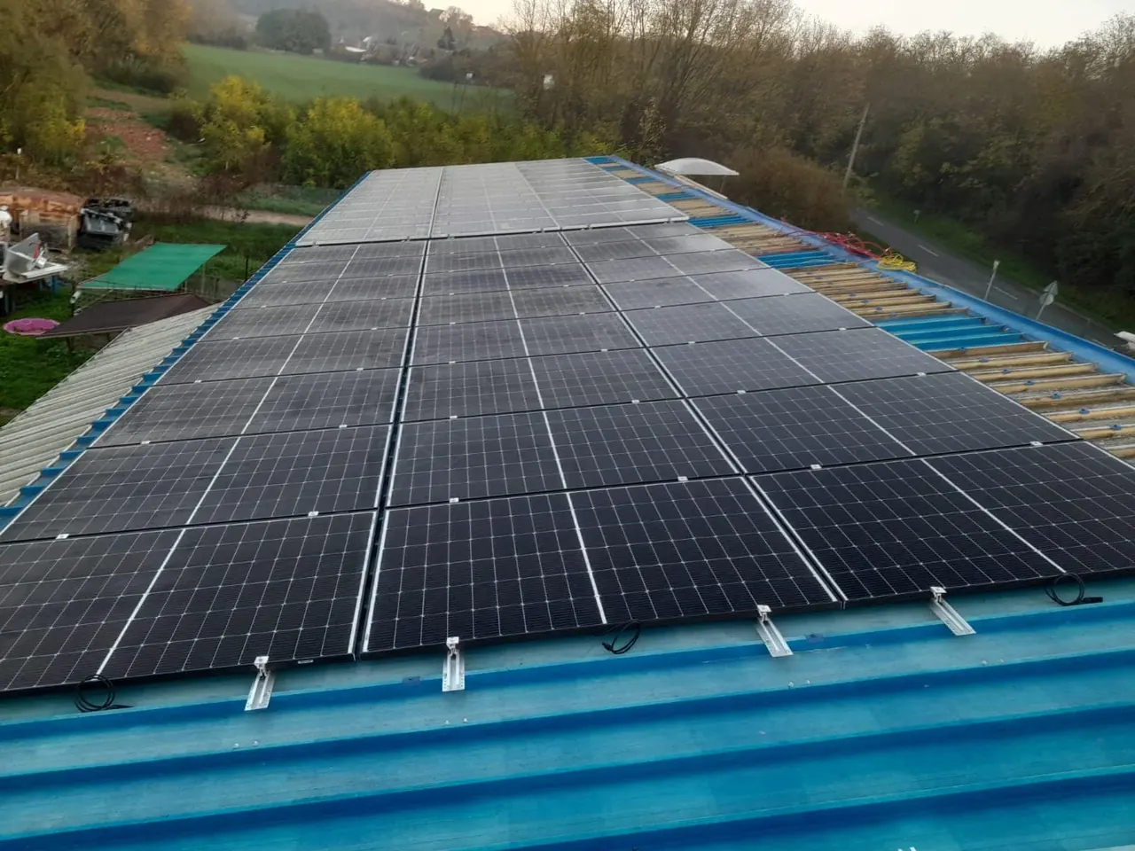 Napelemek sorakoznak egy épület tetején Baján, a Ares Solar Construction Kft. napelem telepítési projektjében. A képen jól látható a napfényes időjárás, amely elősegíti a napelemek hatékony működését. A zöld táj és a fák a háttérben természetes környezetet biztosítanak, tükrözve a fenntartható energia iránti elköteleződést.