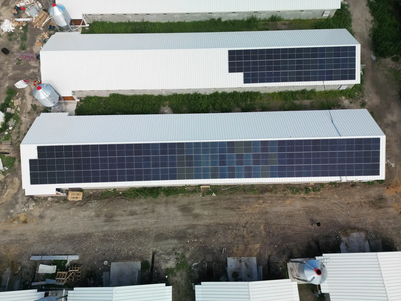 A képen Dócon, az Ares Solar Construction Kft. által végzett napelem telepítés látható. A felvételen két hosszú, fehér tetős épület látható, amelyek tetején sötét napelemek sorakoznak. Az épületek környezetében zöld növényzet és földút található, ami a vidéki táj hangulatát idézi. A napelemek modern technológiát képviselnek, hozzájárulva a fenntartható energiahasználathoz.