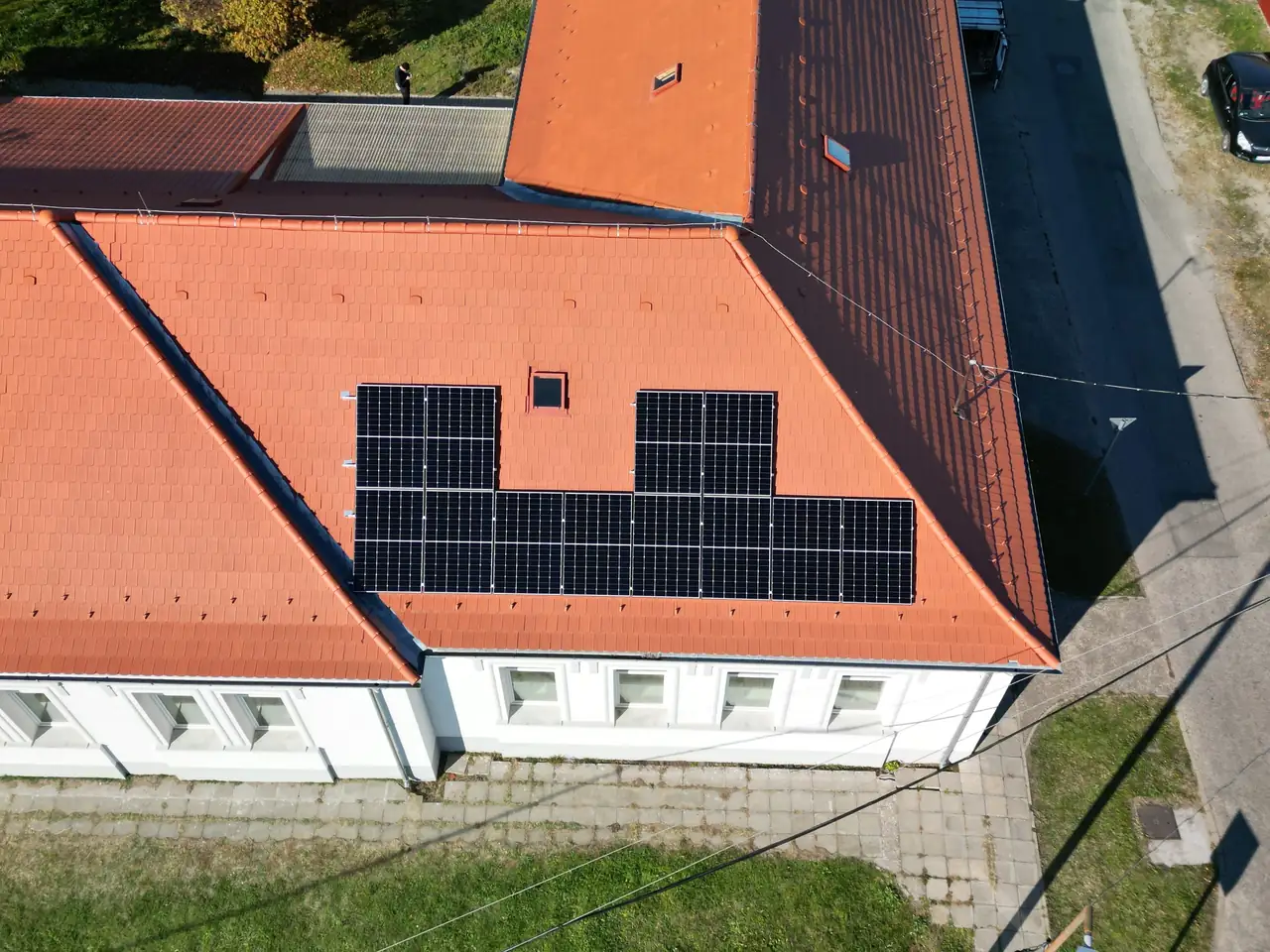 Ezen a képen egy Miskolcon található ház látható, amelynek tetején napelemek vannak telepítve. A piros cseréptető közepén a napelemek rendszere jól látható, ahogy a napfényben csillognak. Az Ares Solar Construction Kft. szakértője által végzett napelem telepítés jól illeszkedik az épülethez, modern megjelenést kölcsönözve annak. A zöld fű és az ingatlan környezete barátságos hátteret biztosít.