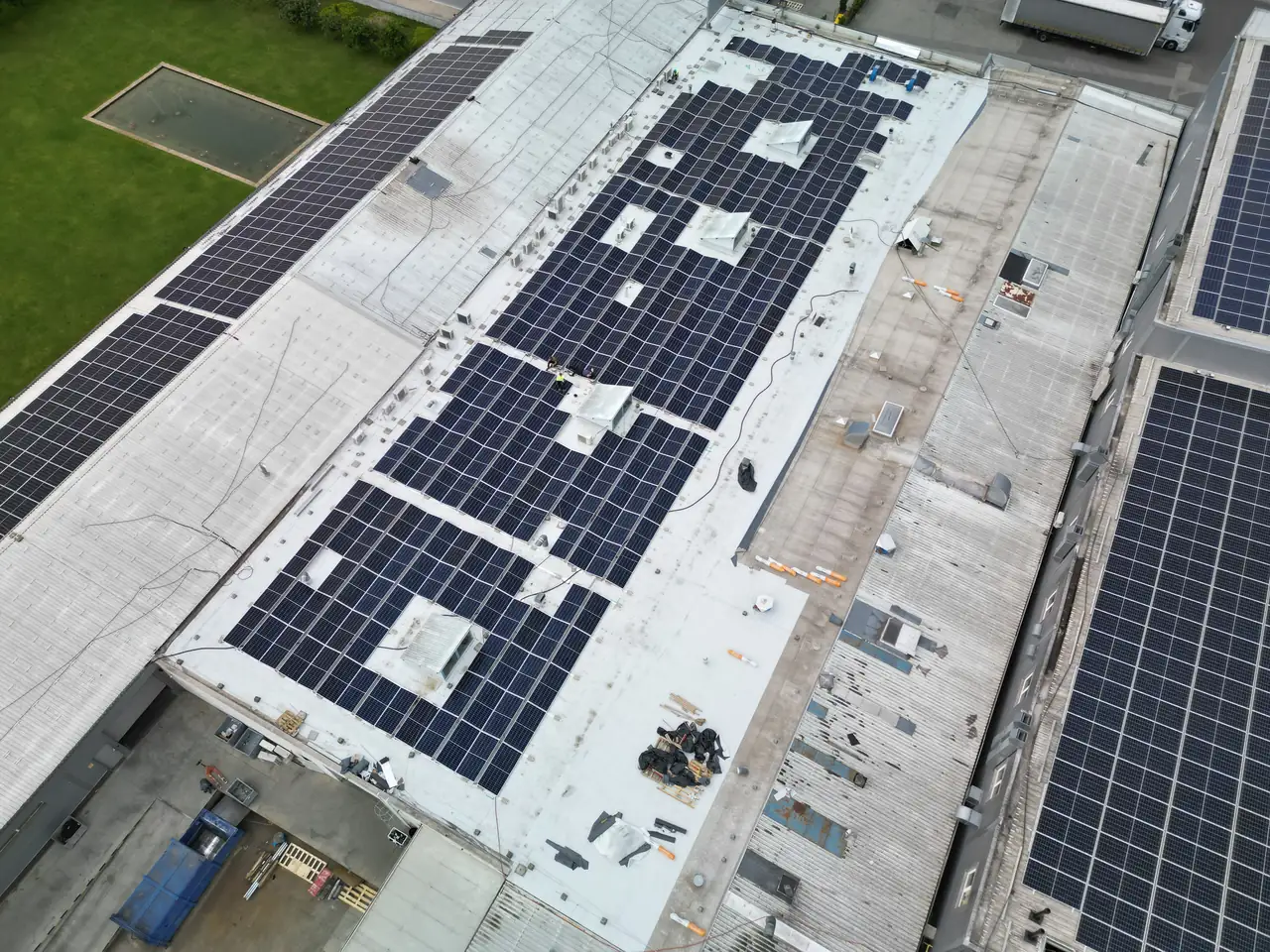 Hajdúszoboszló városában, az Ares Solar Construction Kft. által végzett napelem telepítés látható egy ipari épület tetőjén. A tetőn sűrűn elhelyezett napelemek fényes felületei tükrözik a napfényt, míg az épület körüli zöld terület és a szomszédos járművek részletei is megfigyelhetők. Az installáció professzionális munkát tükröz, amely hozzájárul a fenntartható energiahasználathoz.