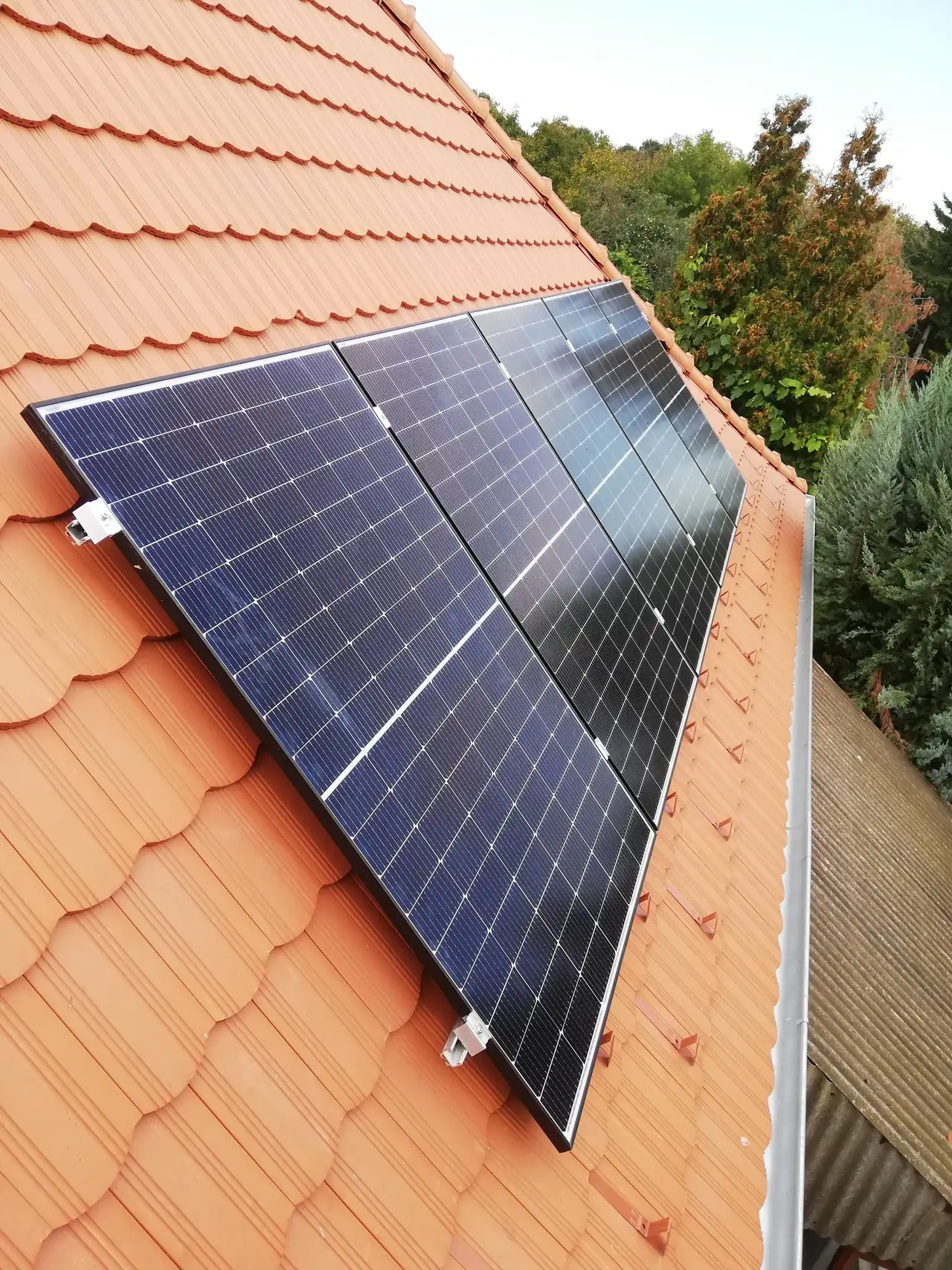 Napelem telepítése Szolnokon, az Ares Solar Construction Kft. által. Az image a tetőn elhelyezett napelemeket mutatja, amelyek hatékonyan hasznosítják a napenergiát. A napelemek sötét színnel és modern dizájnnal rendelkeznek, a cseréptető piros árnyalatai mellett, ami jól látható a háttérben lévő zöld fák mellett.