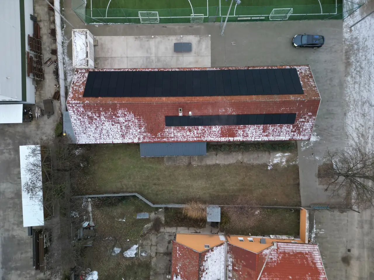 Hódmezővásárhelyen, az Ares Solar Construction Kft. napelem telepítésének eredménye látható egy épület tetején. Az épület vörös cseréppel fedett, amelyen fekete napelemek sorakoznak. Az alatta elhelyezkedő zöld fű és az épület körüli terület jól látható, míg a háttérben sportpályák is találhatók. Az összkép a fenntartható energiaforrások iránti elkötelezettséget tükrözi.
