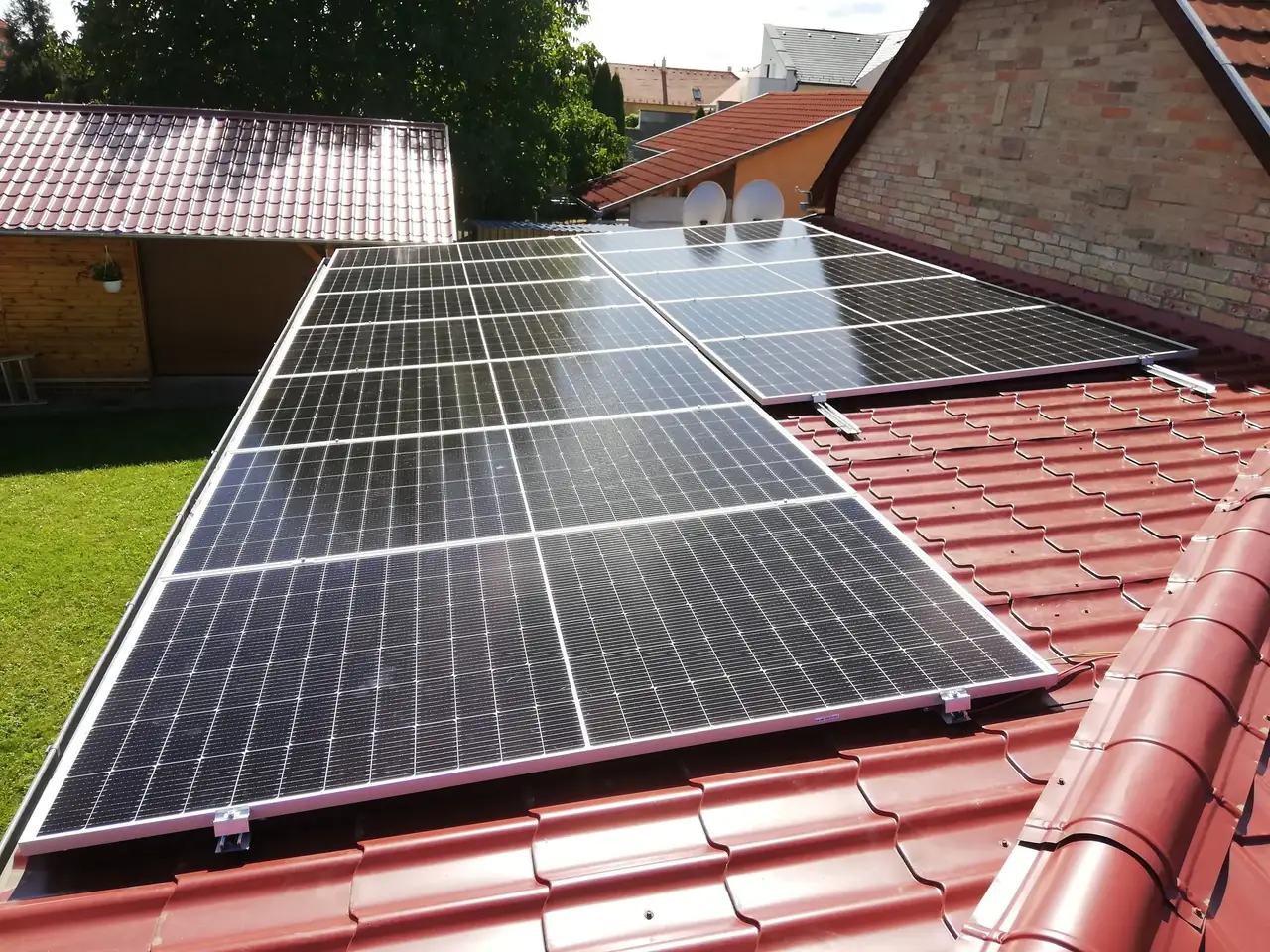 Napelem telepítése Dóc városában, az Ares Solar Construction Kft. által végzett projekt keretében. A képen látható tetőn több napelem található, amelyek a vörös cserépfedésen helyezkednek el. A háttérben lakóépületek és autók láthatók, ami a település jellegzetes látképére utal. A napelemek modern technológiát képviselnek, és a fenntartható energiaforrások hasznosítását szolgálják.