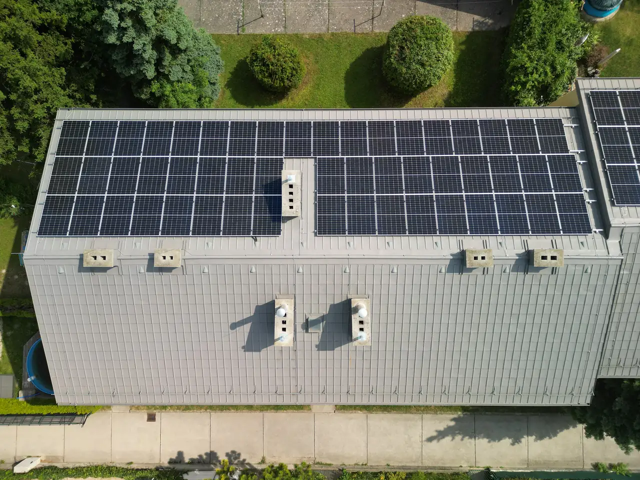 Tatabányán, az Ares Solar Construction Kft. napelem telepítési projektje látható, ahol egy modern épület tetején számos napelem található. A napelemek a napenergia hatékony hasznosítását szolgálják, miközben a zöld környezet harmonikus hátteret biztosít. Az épület körüli zöld területek, valamint a jól megtervezett telepítés a fenntartható energia megoldások iránti elkötelezettséget tükrözik.