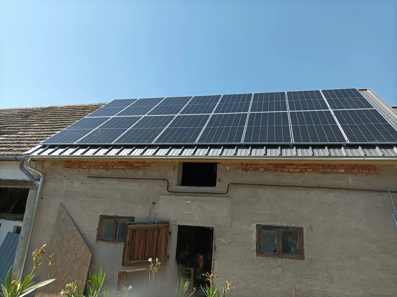 Napelem telepítése Veszprémben, ahol az Ares Solar Construction Kft. szakszerűen helyezte el a napkollektorokat egy ház tetején. A képen jól látható a tetőre szerelt napelem-rendszer, amely a napfény energiáját hasznosítja. A háttérben a ház falának textúrája és ablakai is láthatók, ami a telepítés környezetét mutatja be.