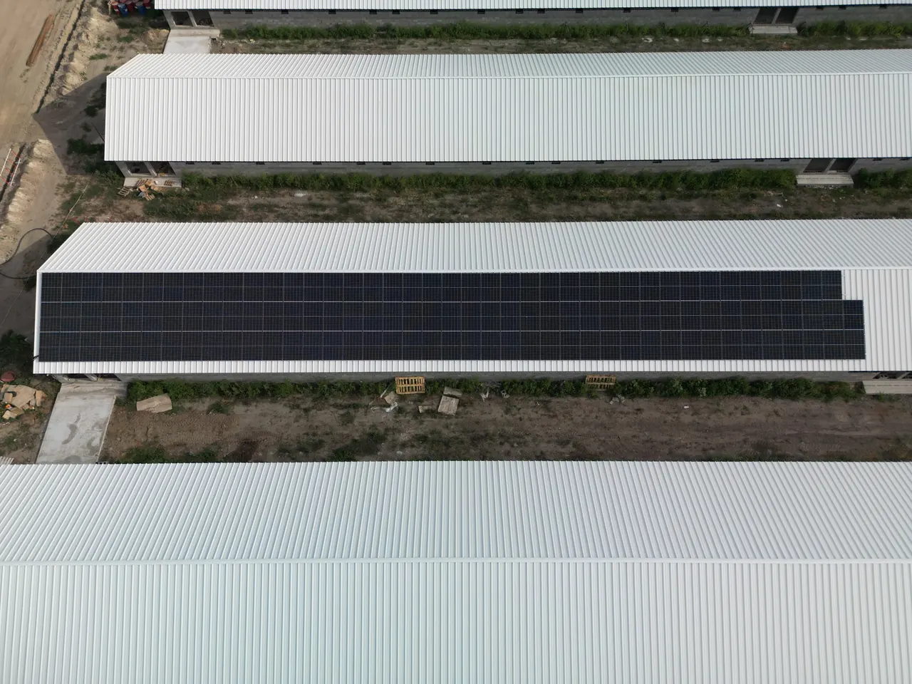 A képen a Dócon található Ares Solar Construction Kft. napelem telepítésének munkálatai láthatók. A tetőkön elhelyezett napelem panelek széles sávban futnak, míg az épületek között zöld területek és építkezési anyagok találhatóak. A modern autótöltő szolgáltatás érdekében készült telepítés kiemelkedő példája a fenntartható energiahasználatnak.