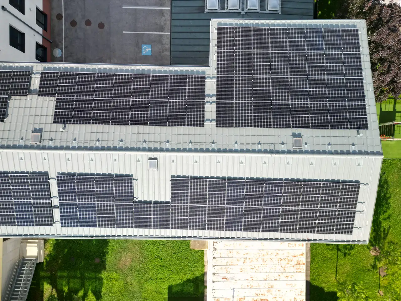 A képen egy modern épület tetején elhelyezett napelemek láthatók, amelyek a Dócon található Ares Solar Construction Kft. napelem telepítési munkálatait tükrözik. Az épület környezetében zöld fű terül el, a háttérben parkoló látható. A napelemek energiatakarékos megoldást kínálnak, ami elősegíti a fenntartható fejlődést a városban.