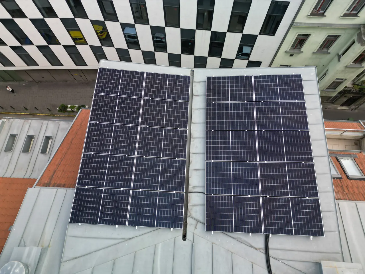 A képen egy modern épület tetején elhelyezett napelemek láthatók, amelyek a Miskolc városában található Ares Solar Construction Kft. Napelem Telepítés szolgáltatásának részeként készültek. A napelemek sötét színűek, és szabályos elrendezésben helyezkednek el, jól láthatóan a tető szélén. Az épület mögött több más városi épület is látszik, amelyek a város urbanizált környezetét tükrözik.