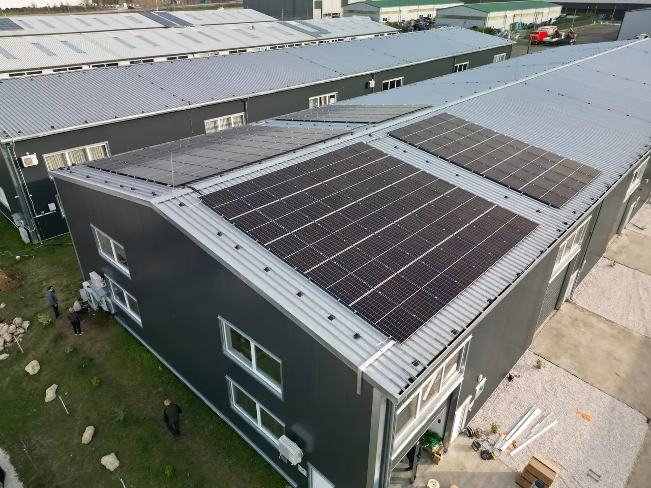 A képen egy modern épület látható Szolnokon, amelynek tetőjén napelemek sorakoznak. Az Ares Solar Construction Kft. által végzett napelem telepítés eredményeként a tető hatékonyan hasznosítja a napenergiát. Az épület körüli zöld területen munkások láthatók, akik a telepítés befejezésén dolgoznak. Az ipari környezetben a napelemek kiemelkedő szerepet játszanak a fenntartható energiahasználatban.