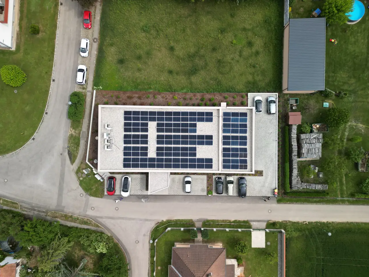 A képen Dóc városában található Ares Solar Construction Kft. napelem telepítési projektje látható madártávlatból. A modern épület tetején több sor napelem panel helyezkedik el, amelyek a napenergiát hasznosítják. A környező területen parkoló autók és zöld területek láthatók, amelyek a telepítés környezetbarát jellegét hangsúlyozzák.