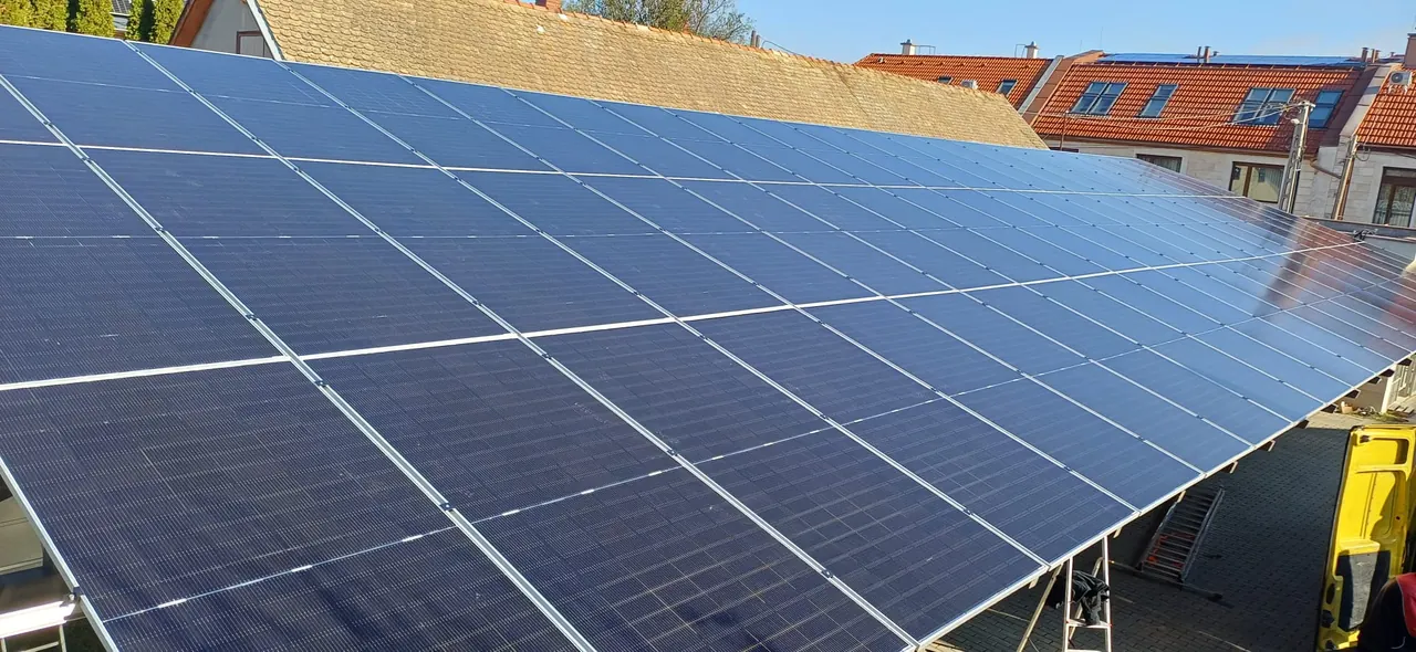 Napelemek sorakoznak egy épület tetején Balaton városában, a Ares Solar Construction Kft. napelem telepítő projektjének keretében. Az éppen napsütötte, fényes napelemek hatékonyan hasznosítják a napenergiát, hozzájárulva a fenntartható energiaforrásokhoz. A zöld energia iránti elkötelezettség jól látható a modern telepítési technológián.