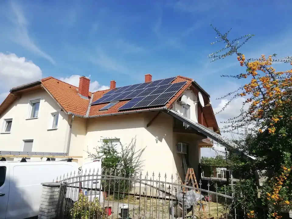 A képen egy Dóc településen található ház látható, amelynek tetőjén napelemek vannak telepítve. A ház fehér színű, piros cserépborítással, és a kertben zöld növények láthatók. A napelem telepítését az Ares Solar Construction Kft. végezte, hangsúlyozva a fenntartható energiaforrások használatát. Az épület mellett egy furgon is parkol, ami a kivitelezéshez szükséges eszközöket szállítja.