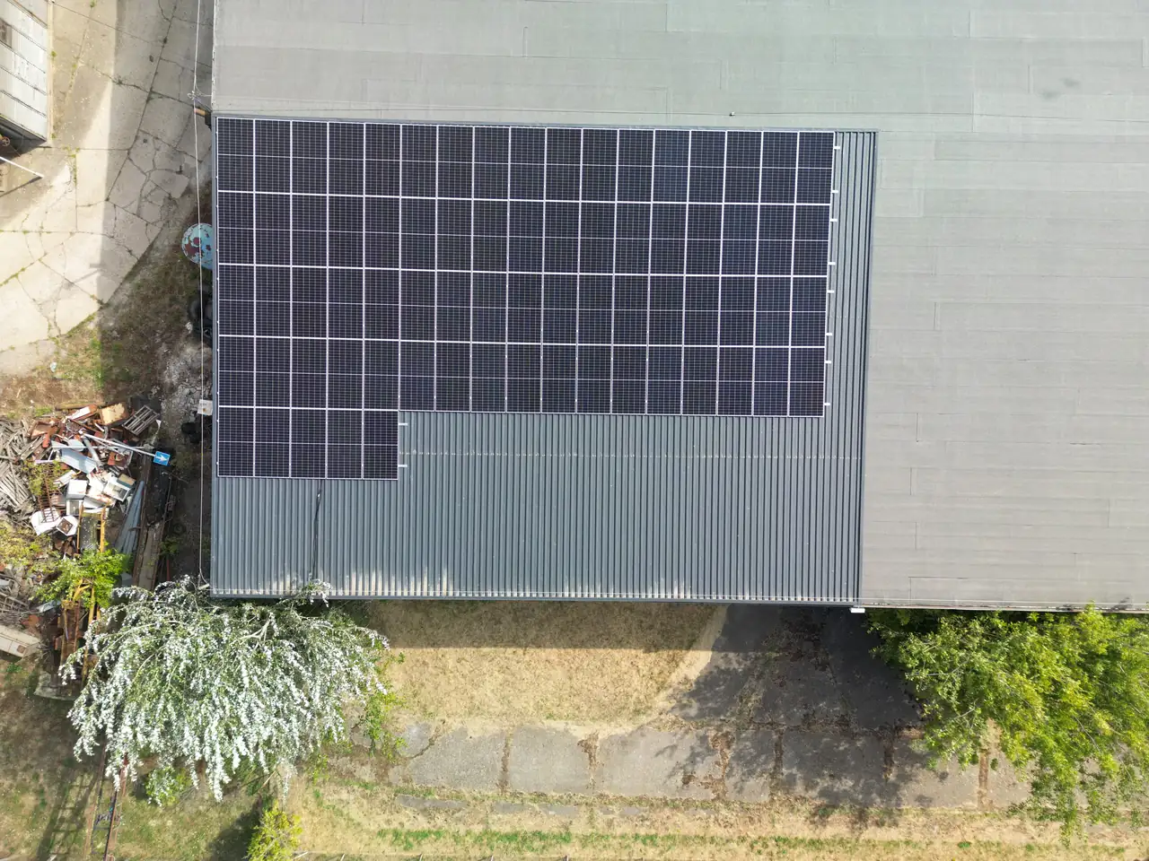 Eger városában, az Ares Solar Construction Kft. által végzett napelem telepítés látható egy ipari épület tetején. A képen jól látszanak a fekete napelemek, amelyek hatékonyan hasznosítják a napenergiát. Az épület körül zöld növényzet és egy szürke aszfaltúton lévő terület is látható, amely a fenntartható energiaforrások iránti elkötelezettséget tükrözi.