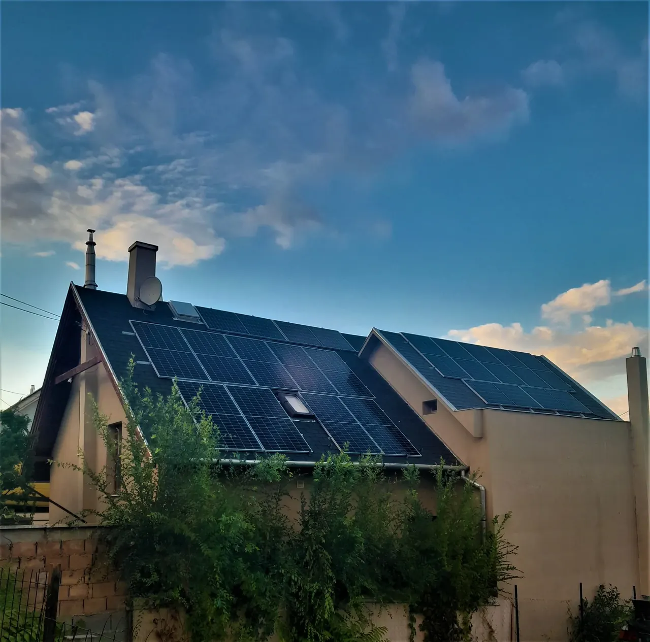 A képen egy családi ház látható, amelynek tetején napelemek sorakoznak. A napelemek a Dócon található Ares Solar Construction Kft. által végzett napelem telepítés eredményeként kerültek felszerelésre. A háttérben gyönyörű kék ég és felhők láthatók, míg a ház körül zöld növényzet borítja a területet.