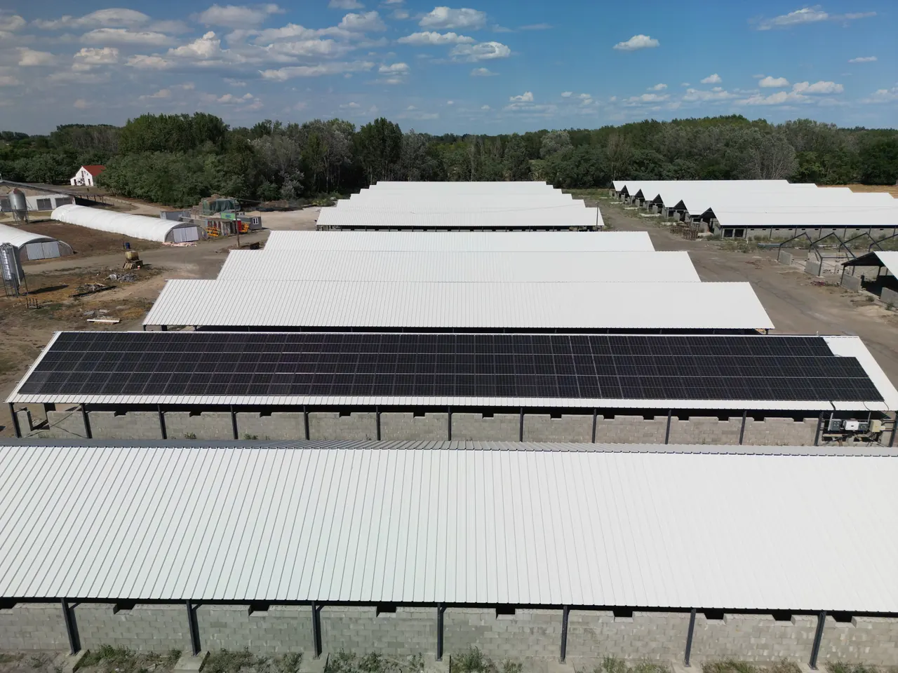 A képen a Dócon található Ares Solar Construction Kft. napelem telepítésének munkálatai láthatók. Az épületek tetején fekvő napelemek szépen kirajzolják a területet, miközben a háttérben zöld fák és kék égbolt található. Az ipari létesítmény modern megjelenése és a napenergia kihasználása jól illeszkedik a fenntartható fejlődéshez.