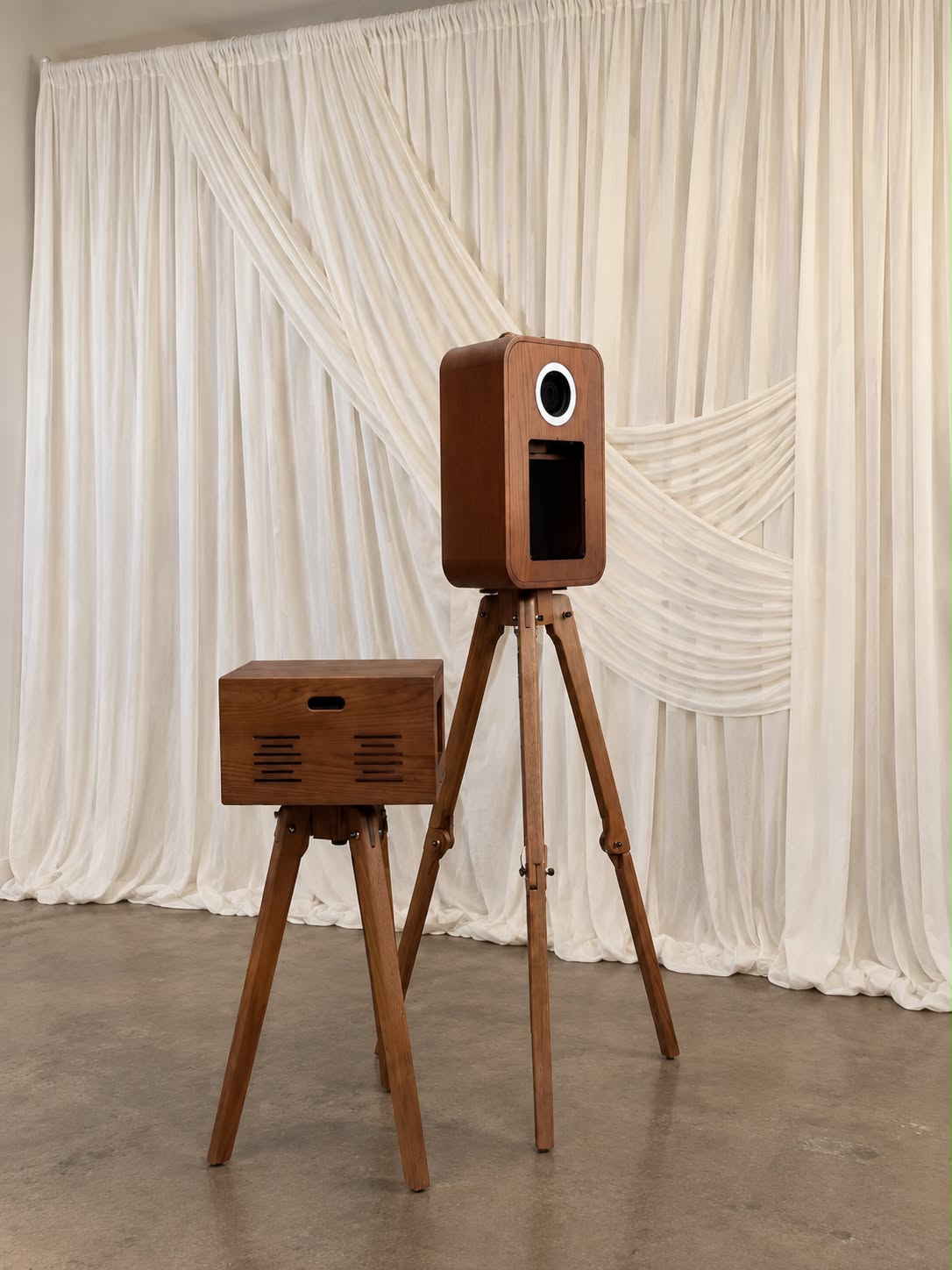 Vintage-style wooden photo booth camera on a tripod beside a matching wooden print stand, set against soft ivory draped curtains