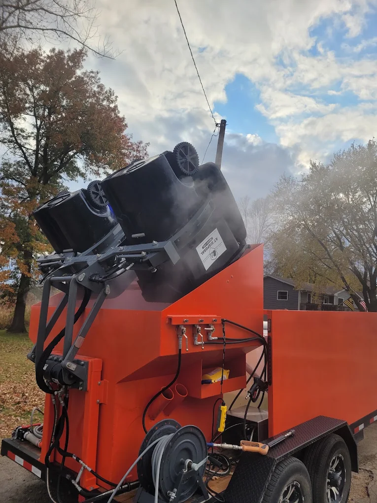 An orange industrial trailer with a waste bin cleaning system lifting two black bins emitting steam on a cloudy day.