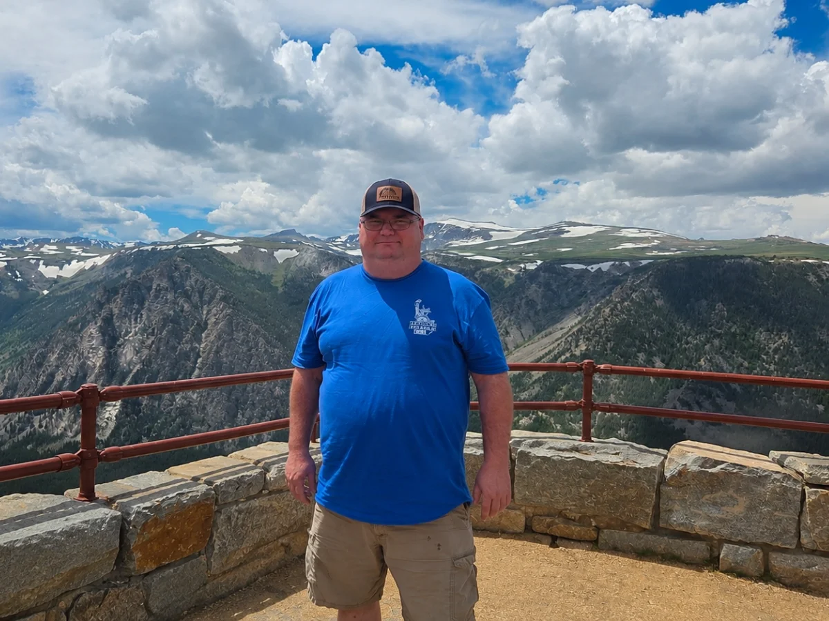 A man in a blue shirt and baseball cap stands on a stone overlook with mountains and snowy peaks in the background.