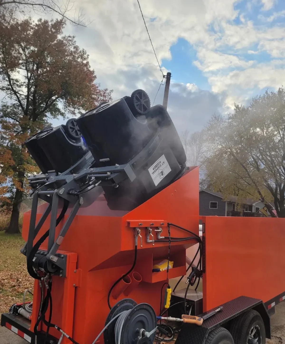 Two black garbage bins upside down on an orange cleaning machine, emitting steam against a cloudy sky.
