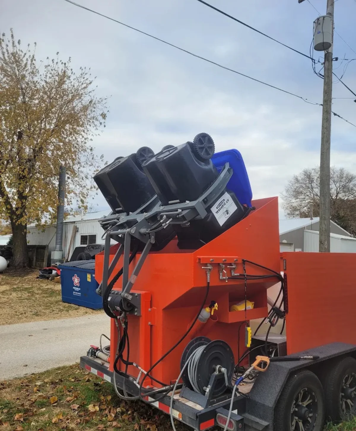 A bright orange trailer with an automated lift system for garbage bins, featuring three bins loaded and ready to be emptied.