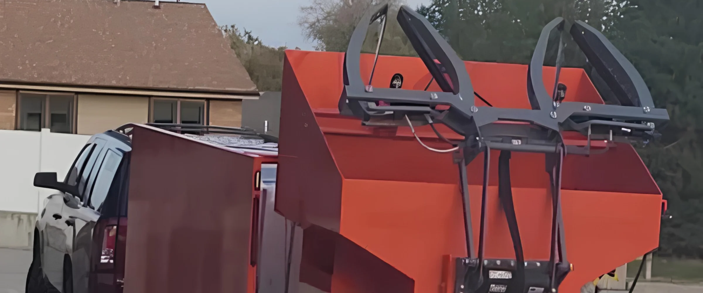 A bright orange dumpster truck trailer with dark gray claw attachments, parked next to a silver pickup truck.
