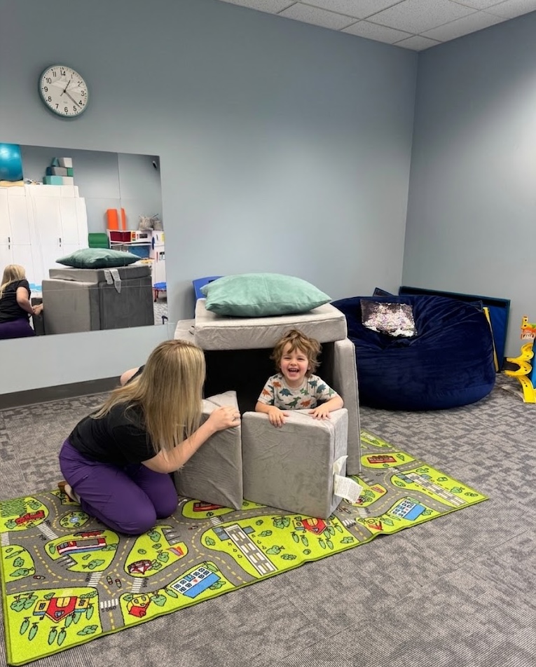Child laughing inside a cushion fort while SLP plays alongside