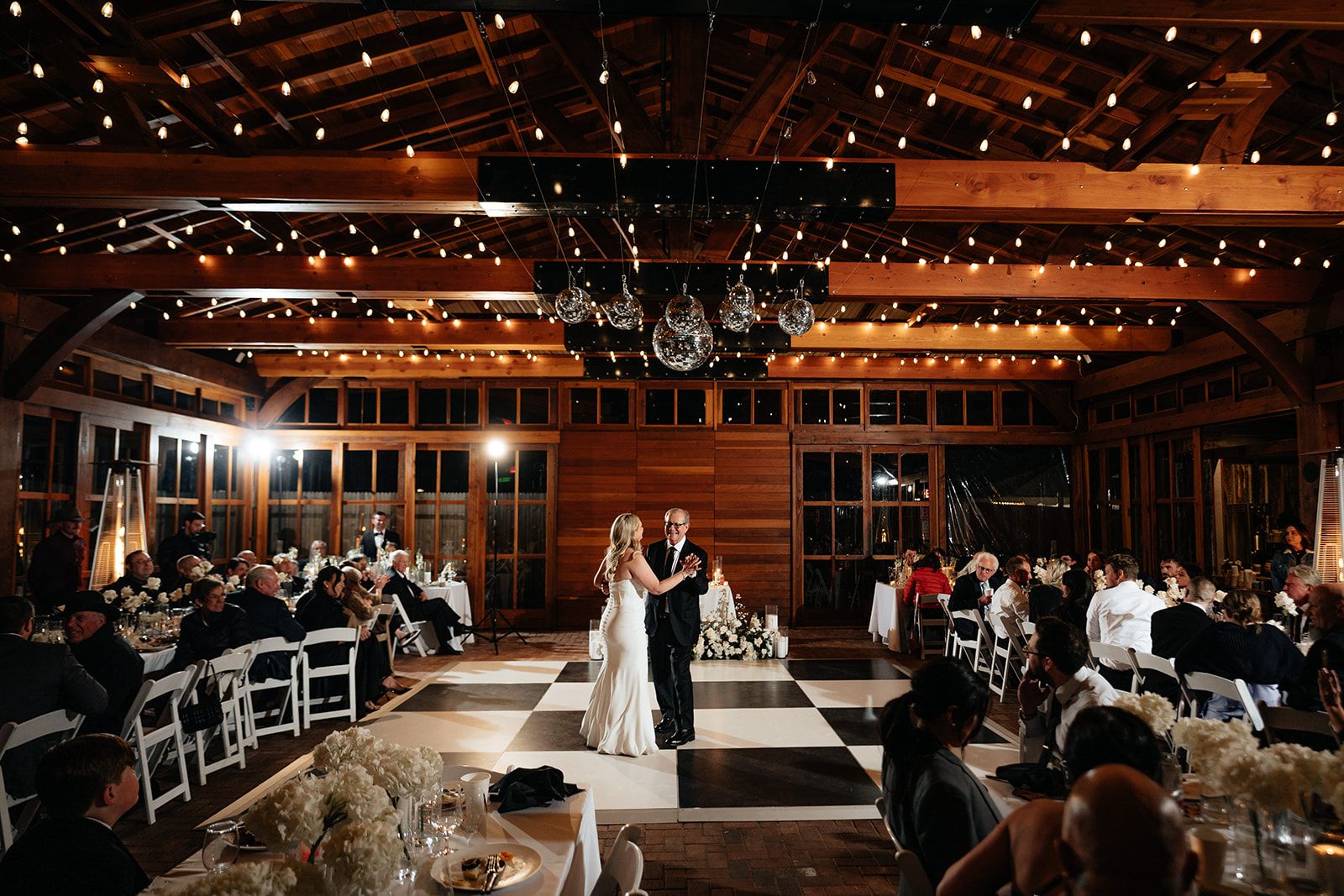 A bride dances in a timber lodge reception under string lights and mirror balls while guests watch