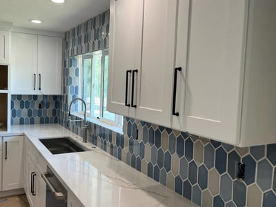 Modern kitchen with a blue and white hexagonal tile backsplash installed behind white cabinetry and a large window, demonstrating high-quality kitchen tile work.