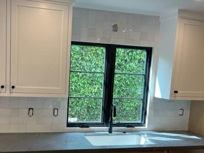 Kitchen with a simple white square tile backsplash surrounding a large window, complemented by dark black window frames and modern cabinetry.