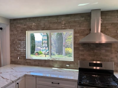 Kitchen with a rustic brown subway tile backsplash and stainless steel range hood, designed to complement white cabinetry and a large window.