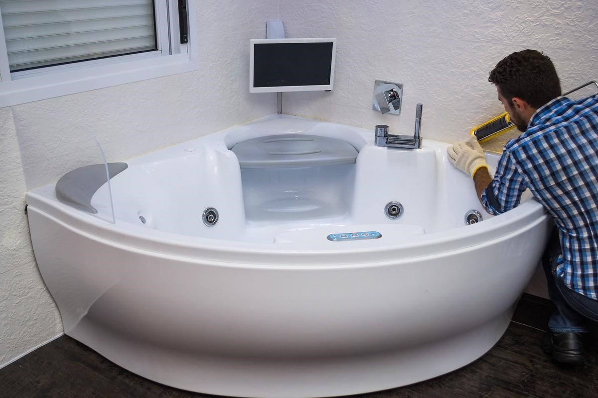 A plumber applying silicone caulk around a white corner whirlpool jacuzzi tub with a small built-in TV screen.