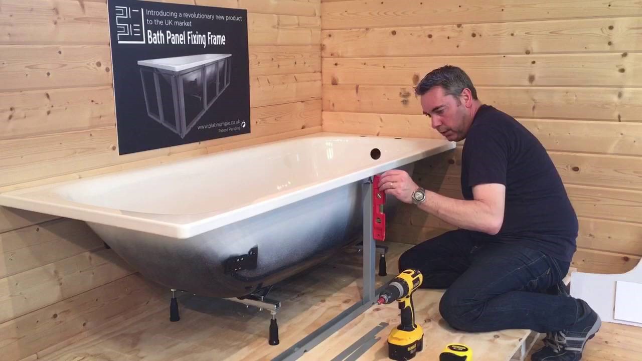 Man installing a Bath Panel Fixing Frame on a white acrylic bathtub using a spirit level and cordless drill.