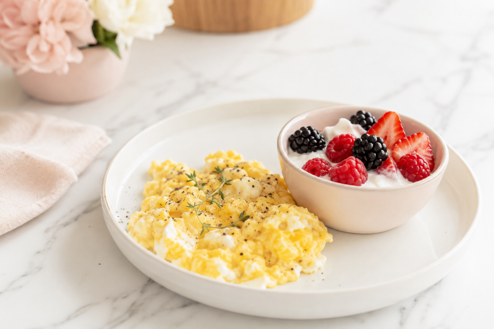 Soft scrambled eggs and a bowl of Greek yogurt topped with berries on a white marble counter. Soft scrambled eggs and a bowl of Greek yogurt topped with berries on a white marble counter.